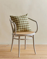 A white wooden chair sits on a wooden floor with the Hawkins New York Essential Gingham Pillow in Sage/Olive (18x18 inches), featuring a plush down insert, set against a neutral background.
