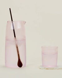 A Hawkins New York Essential Glassware - Pitcher in Blush stands beside a matching pink glass, both filled with water and accompanied by a long metal spoon, all set against a plain background.