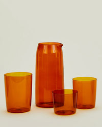 A Hawkins New York Essential Glassware - Pitcher in Terracotta sits next to three matching cups of varying sizes, all arranged against a plain background.