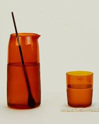 A Hawkins New York Essential Glassware - Pitcher in Terracotta, with a stir stick inside, stands beside a matching amber glass cup of water on a white surface.