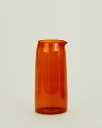 The Hawkins New York Essential Glassware Pitcher in Terracotta, crafted from translucent orange borosilicate glass and featuring a spout, is centered against a plain off-white background.