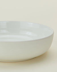 A close-up of a plain, bone-colored ceramic bowl from the Essential Low Bowls in Bone - Set of 4 by Hawkins New York, shown on a light surface.