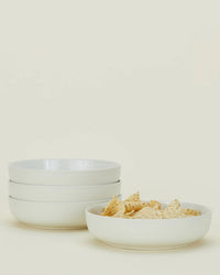 Four Hawkins New York Essential Low Bowls in Bone are stacked on the left, with one matching bowl filled with tortilla chips on the right—versatile stoneware dinnerware ideal for any occasion, set against a simple light background.