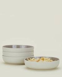 Four stacked Essential Low Bowls in Light Grey by Hawkins New York sit beside a shallow bowl of tortilla chips, all set against a plain light background.