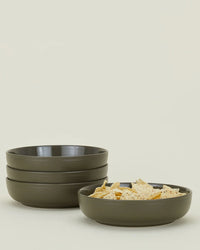 Four olive Essential Low Bowls by Hawkins New York are stacked, with a fifth matching bowl in front holding tortilla chips. This stylish stoneware set of 4 is displayed against a plain light background.