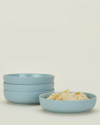 A set of four Essential Low Bowls in Sky by Hawkins New York is stacked beside a matching bowl filled with crackers, all crafted from stoneware and displayed against a plain light background.