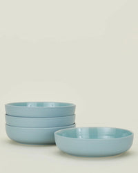 Four Hawkins New York Essential Low Bowls in Sky are stacked, with a fifth bowl in front, highlighting the elegant stoneware set against a simple background.
