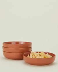 Four stacked Hawkins New York Essential Low Bowls in Terracotta (Set of 4) sit beside a matching bowl of tortilla chips, highlighting the elegant stoneware dinnerware against a plain light background.
