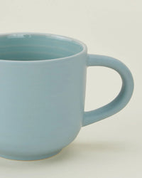 A close-up of a Hawkins New York Essential Mug in Sky, featuring a handle and light blue finish—part of their stoneware Essential Mugs in Sky set of 4.