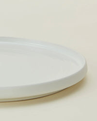 A close-up of the edge of a plain, round, white ceramic plate from the Hawkins New York Essential Salad Plates in Bone (Set of 4) on a light-colored surface.