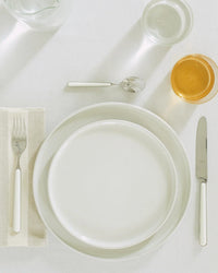 A neatly set dining table features Hawkins New York Essential Salad Plates in Bone (Set of 4) stacked on a white tablecloth, with a fork, knife, spoon, beige napkin, water glass, and a glass of amber liquid.