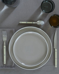 A fork, knife, spoon, gray napkin, and two dark glass tumblers are arranged beside stacked Hawkins New York Essential Salad Plates in Light Grey (Set of 4) on a gray tablecloth.