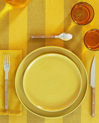 A yellow table setting features stacked Hawkins New York Essential Salad Plates in Mustard, wooden-handled fork, knife, and spoon, a napkin, and two amber glasses on a yellow striped stoneware dinnerware tablecloth.