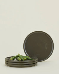 Four Hawkins New York Essential Salad Plates in Olive are stacked, topped with three pieces of okra; one plate stands upright behind the stack against a plain background.