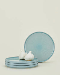 Four Hawkins New York Essential Salad Plates in Sky are stacked with three garlic bulbs on top, and one plate is propped upright behind them against a plain, light backdrop that highlights the elegant stoneware.