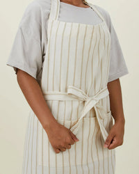 Someone wears the Hawkins New York Essential Striped Apron in Ivory/Flax—crafted in cotton with vertical stripes, front pockets, and a tied waist—over a short-sleeved shirt, making it an ideal kitchen gift.