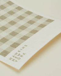 A close-up of Hawkins New York’s Essential Swedish Dishcloths in Ivory/Flax (set of 6), featuring a beige and white check pattern with the brands name printed in the corner.
