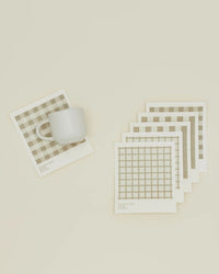 A white ceramic mug sits on an Essential Swedish Dishcloth by Hawkins New York in Ivory/Flax, with five more grid-patterned dishcloths from the set of 6 arranged nearby on a cream surface.