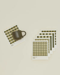 A dark ceramic mug sits on a square Hawkins New York Essential Swedish Dishcloth in Olive/Sage, with four matching dishcloths arranged neatly nearby; the checkered set includes six eco-friendly cloths.
