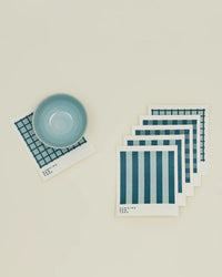 A small blue bowl rests on a grid-patterned napkin beside six Essential Swedish Dishcloths in Sky/Peacock by Hawkins New York, featuring stripe and grid patterns, all arranged on a beige surface.