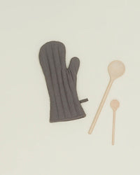A Hawkins New York Simple Linen Oven Mitt in Dark Grey with heat-resistant padding is placed beside two wooden spoons, one large and one small, on a plain light background.