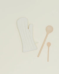 An ivory Simple Linen Oven Mitt by Hawkins New York, featuring heat-resistant padding, is displayed with one large and one small wooden spoon on a plain, light-colored surface.
