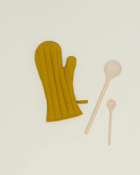 The Hawkins New York Simple Linen Oven Mitt in Mustard sits beside two differently sized wooden spoons on a light cream background.