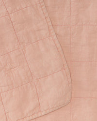 Close-up of the Simple Linen Quilt in Blush by Hawkins New York, featuring a light pink European linen fabric with a grid pattern and stitched details, with one corner of the quilt folded over.