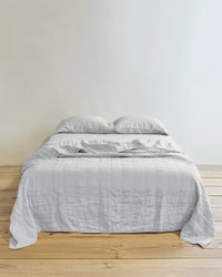 A neatly made bed features the Hawkins New York Simple Linen Quilt in Light Grey and two pillows, resting on a wooden floor against a plain white wall.