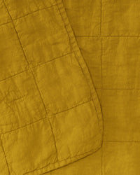 The Simple Linen Quilt in Mustard by Hawkins New York features European linen, visible square-pattern stitching, an overlapping corner, and a subtle stonewashed texture.