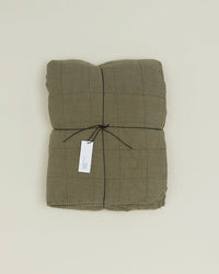 The Hawkins New York Simple Linen Quilt in Olive, featuring a grid pattern, is folded and tied with black string and a white tag, displayed on a plain light background.