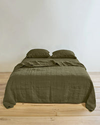 A made double bed featuring the Hawkins New York Simple Linen Quilt in Olive, two pillows, a plain white wall in the background, and a light wooden floor.