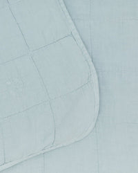 The Simple Linen Quilt in Sky by Hawkins New York features light blue stonewashed linen, a grid stitch pattern, rounded edges, and a small embroidered logo on the corner—offering timeless style for any room.