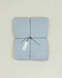 The Hawkins New York Simple Linen Quilt in Sky, neatly folded and tied with black string, features a white tag and rests on a plain light background.