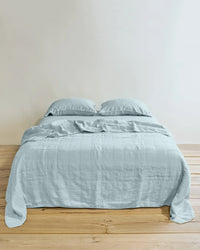 A neatly made bed features the Hawkins New York Simple Linen Quilt in Sky with two pillows, set against a plain off-white wall on a wooden floor.