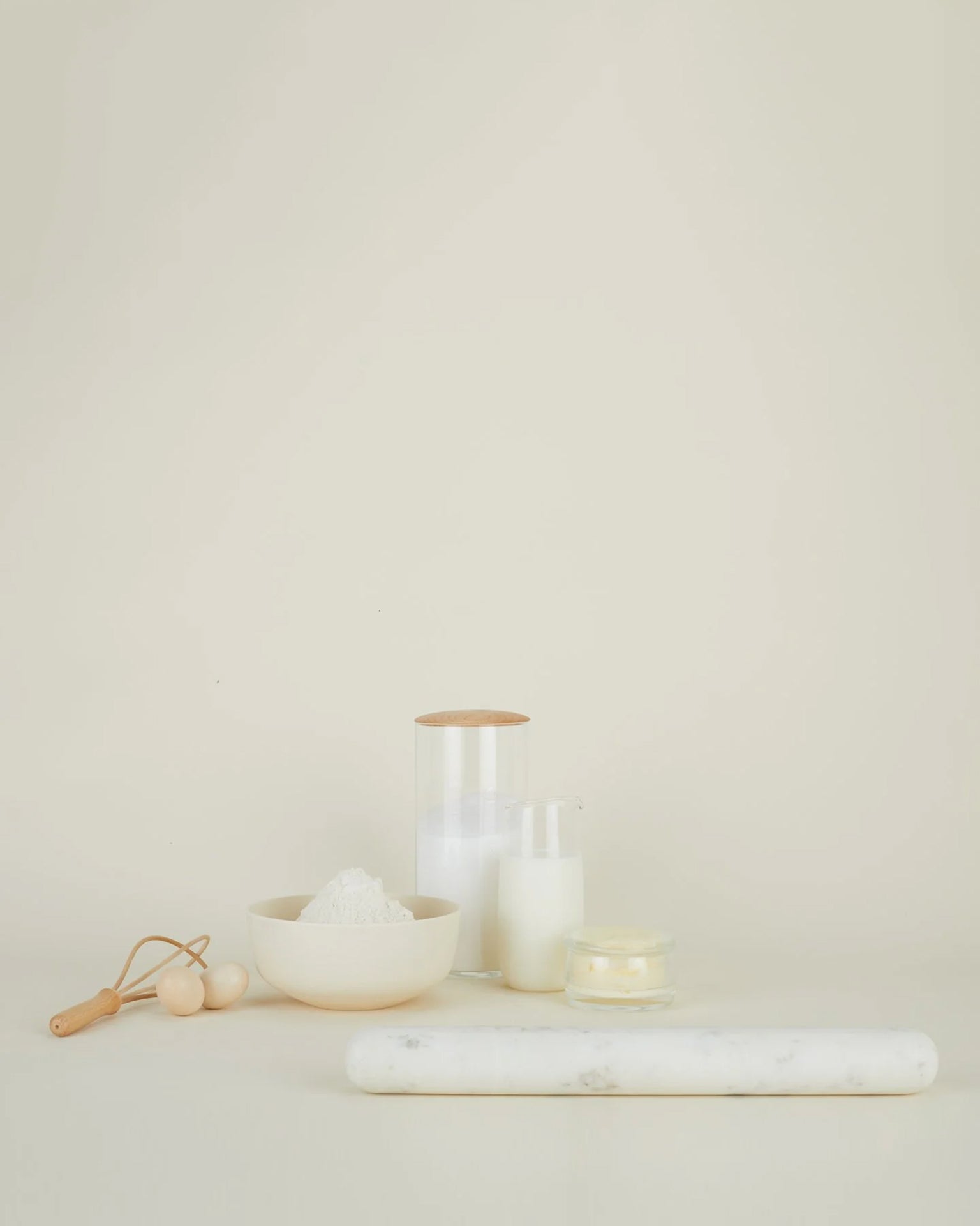 A minimalistic scene with a Hawkins New York Simple Marble Rolling Pin, a bowl of flour, a small glass jar of milk, a lit candle, a storage jar, and a wooden-handled dough cutter on a light beige background.