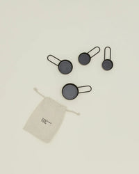 Four black Hawkins New York Simple Measuring Cups are displayed above a small drawstring pouch with the brands label on an off-white background.