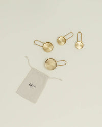 Four brass measuring cups by Hawkins New York are displayed beside a small beige drawstring pouch labeled “YIELDING HOPE,” echoing the elegant style of the brands Simple Measuring Cups.