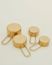 Four Simple Measuring Cups in Brass by Hawkins New York, labeled 1 cup, 1/2 cup, 1/3 cup, and 1/4 cup, are arranged on a light background.