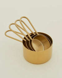 A set of four Hawkins New York Simple Measuring Cups in Brass, each featuring a rounded handle and stacked together on a neutral background. Made from durable stainless steel for lasting elegance and functionality.