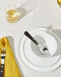 A table setting with a Hawkins New York Simple Porcelain Dinner Plate, matching bowl, a spoon in the bowl, a fork on a yellow napkin, and a glass of light-colored drink atop a crisp white tablecloth.