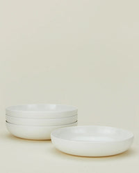 Four plain white shallow bowls from the Hawkins New York Simple Porcelain Dinnerware - 16 Piece Set, three stacked on the left and one placed separately on the right, set against a light beige background.