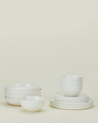 The Hawkins New York Simple Porcelain Dinnerware 16 Piece Set features neatly stacked plates and bowls, all in classic white, elegantly arranged against a light beige background.