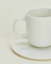 A Simple Porcelain Espresso Cup & Saucer from Hawkins New York sits empty and slightly off-center, its handle on the left, against a light cream background—showcasing minimalist elegance from this stylish set of 2.
