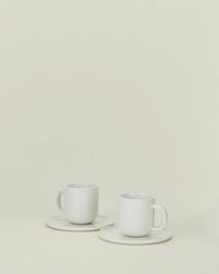 Two Simple Porcelain Espresso Cups & Saucers by Hawkins New York rest on a neutral background, their minimalist design and fine porcelain elegance enhanced by the surrounding empty space. Set includes 2 cups and saucers.