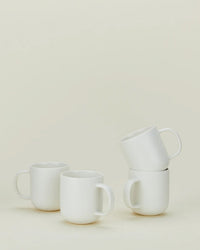 Four Simple Porcelain Mugs by Hawkins New York are arranged on a soft beige surface: two placed side by side, and two stacked with one resting diagonally atop the other.
