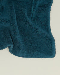A close-up of the Simple Terry Bath Mat in Peacock by Hawkins New York, featuring dark teal textured cotton with curved edges, spread out on a light surface.