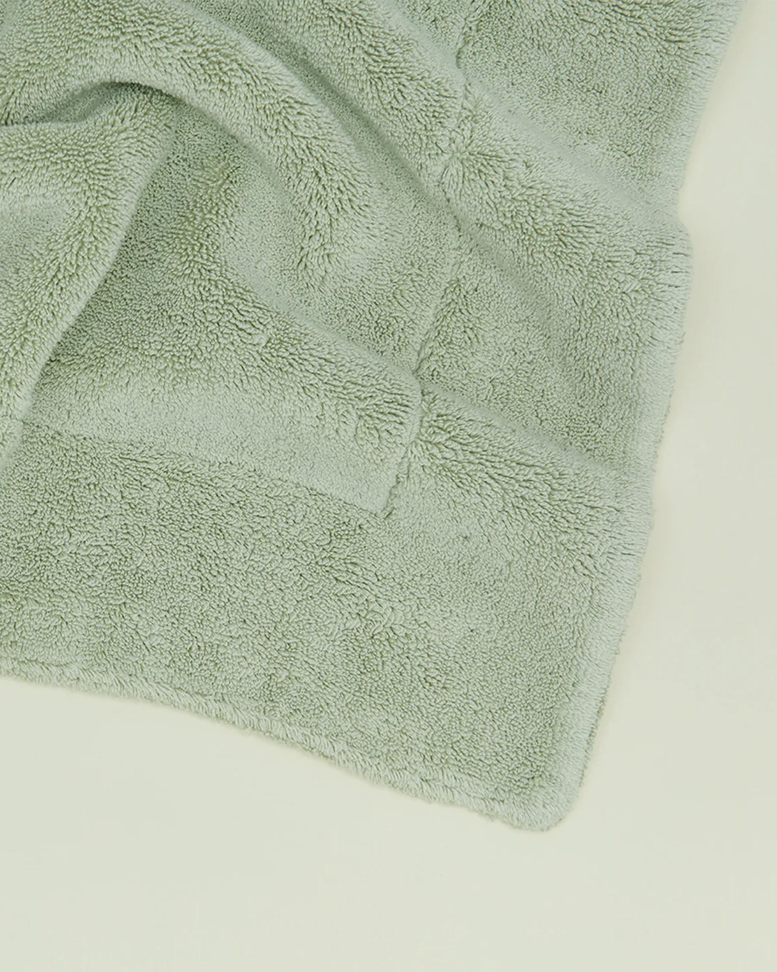 A close-up of a folded, light green, fluffy cotton towel on a pale surface evokes the plush feel of the Simple Terry Bath Mat in Sage by Hawkins New York.