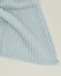 The Simple Waffle Bath Sheet in Sky by Hawkins New York, made from textured blue waffle-knit fabric, is displayed draped over a cream surface, evoking the look of elegant stonewashed cotton towels.