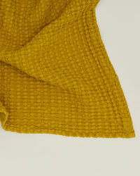 A close-up of the Hawkins New York Simple Waffle Bath Towel in Mustard, made from soft stonewashed cotton with a textured waffle pattern, shown spread out on a plain, light surface.